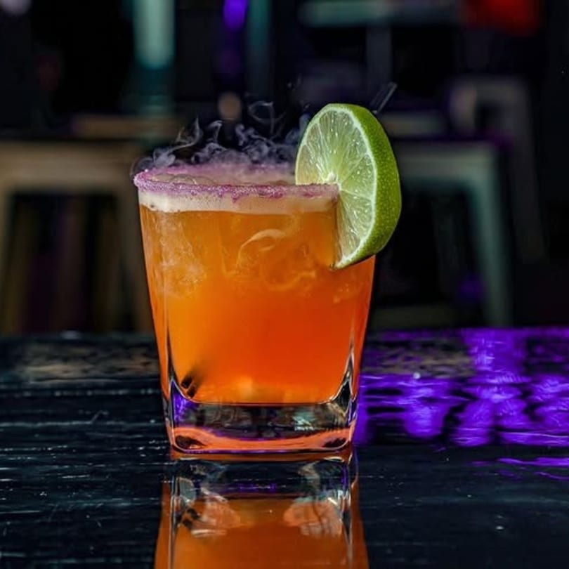 A vibrant orange cocktail with a purple sugar rim, lime wedge, and smoky effect on a dark bar.