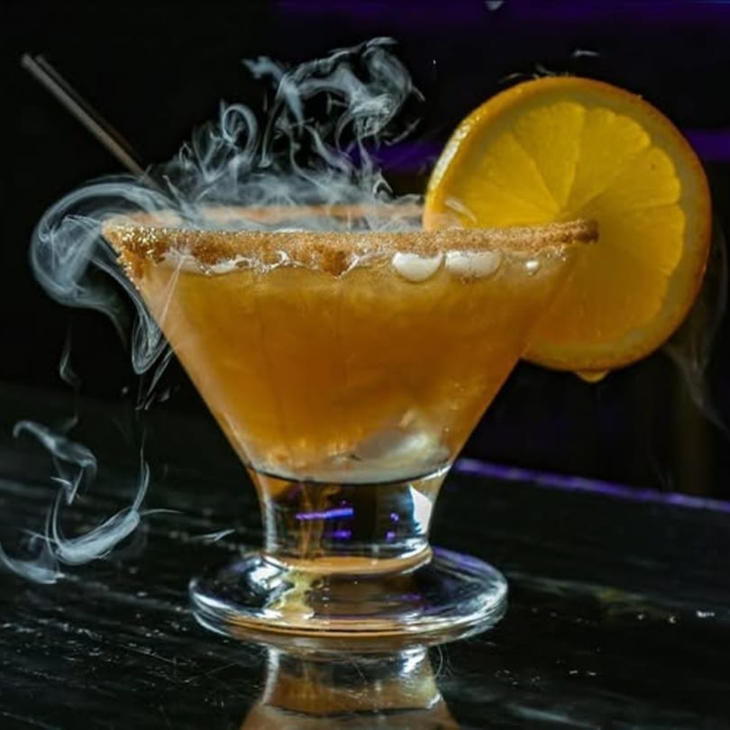 A smoking cocktail with a sugared rim and orange slice, on a dark, reflective surface.