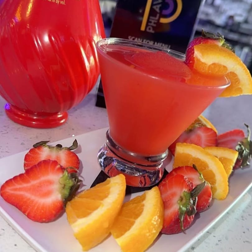 A vibrant red cocktail in a martini glass, garnished with fresh strawberries and orange slices.