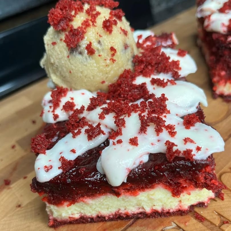 Red velvet cheesecake with white icing, red crumbs, and a scoop of ice cream on a wooden board.