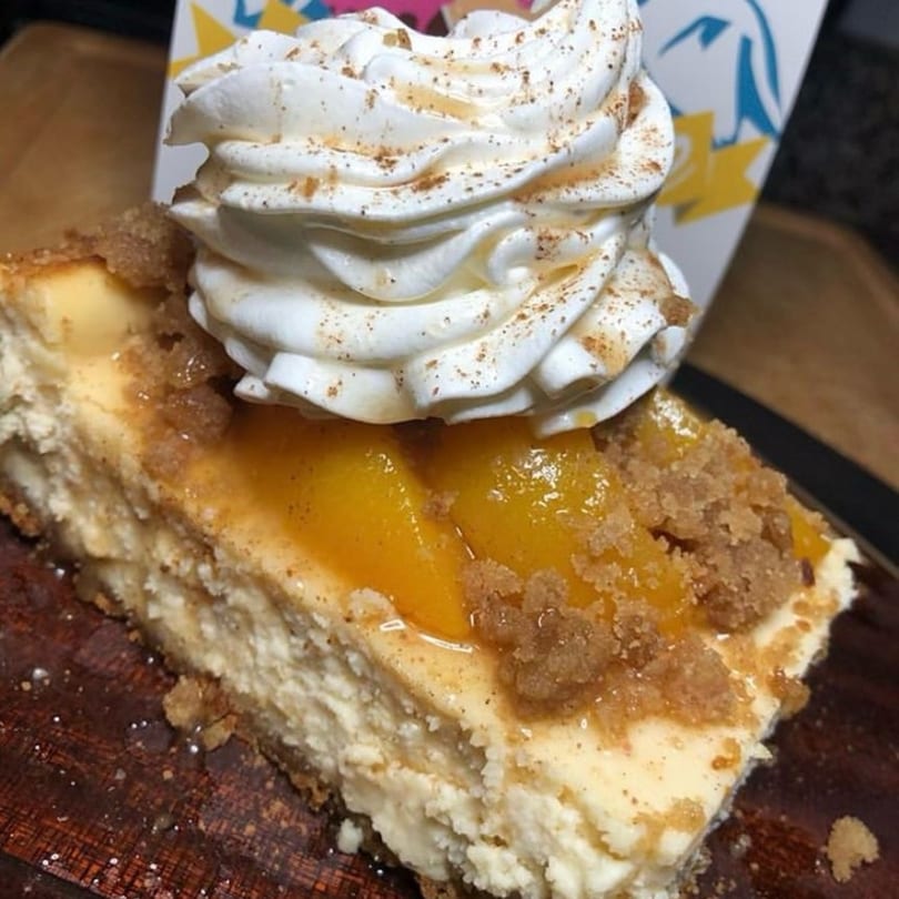 A slice of cheesecake topped with peaches, brown sugar crumble, and whipped cream on a wooden board.
