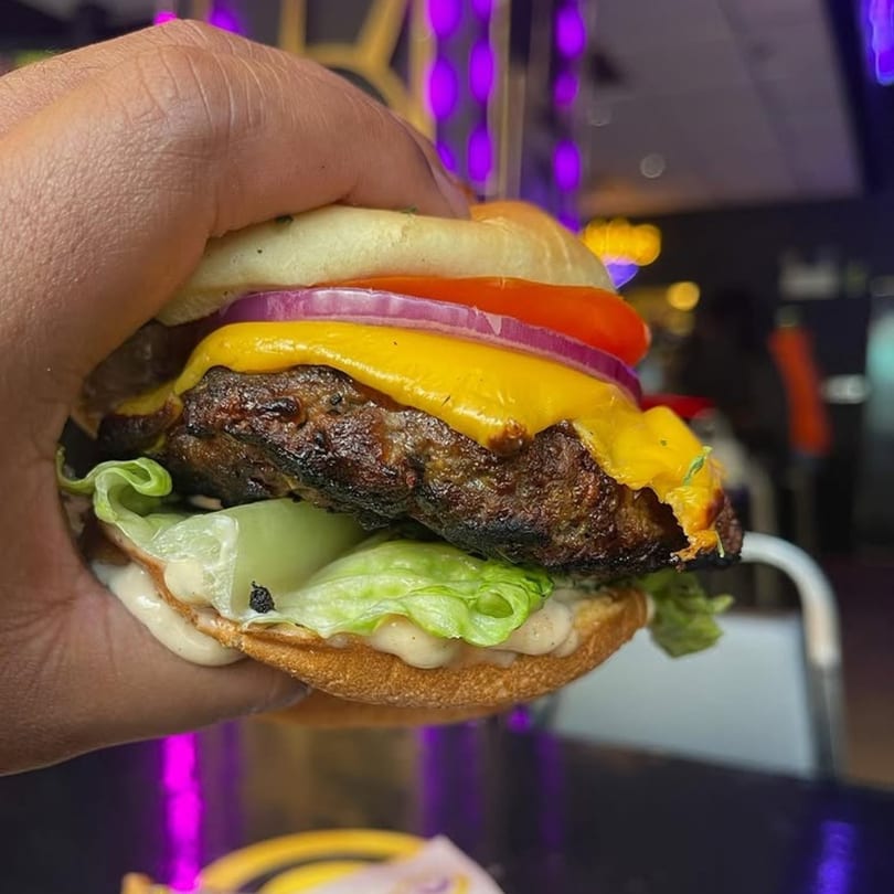 Hand holding a Phlavz burger with melted cheese, lettuce, tomato, and red onion.
